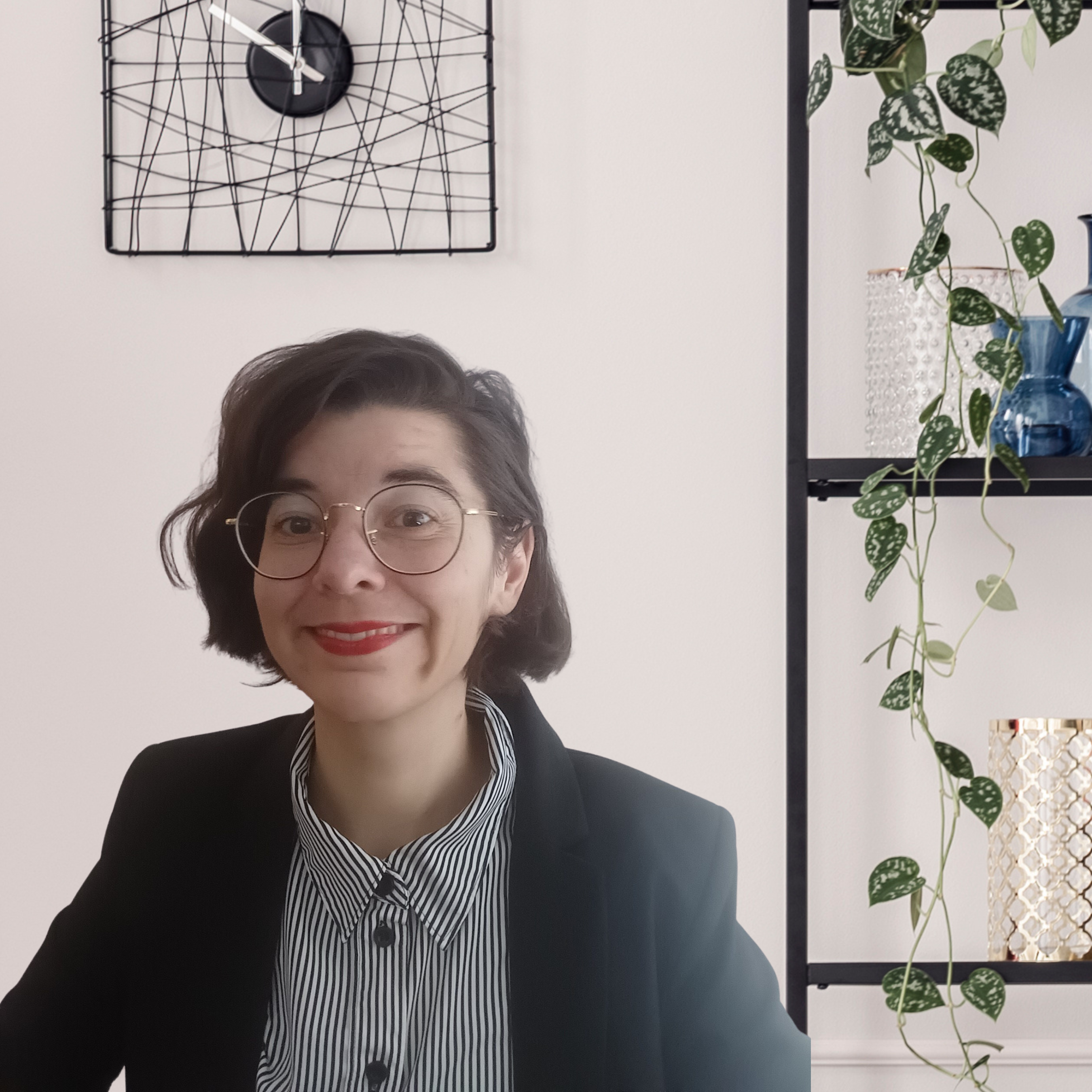Das Bild zeigt eine Person, die vor einem modernen, minimalistischen Hintergrund sitzt. Die Person hat kurzes, dunkelbraunes Haar, trägt eine runde Brille und lächelt freundlich in die Kamera. Sie ist formell gekleidet: in einem schwarzen Blazer und einer gestreiften Bluse mit schwarz-weißen Streifen. Im Hintergrund ist eine stilvolle Uhr mit geometrischem Design an der Wand zu sehen. Rechts davon steht ein Regal, das mit einigen dekorativen Elementen wie einer hängenden Pflanze und Vasen in verschiedenen Farben und Texturen geschmückt ist. Die gesamte Szene wirkt hell, modern und einladend.
